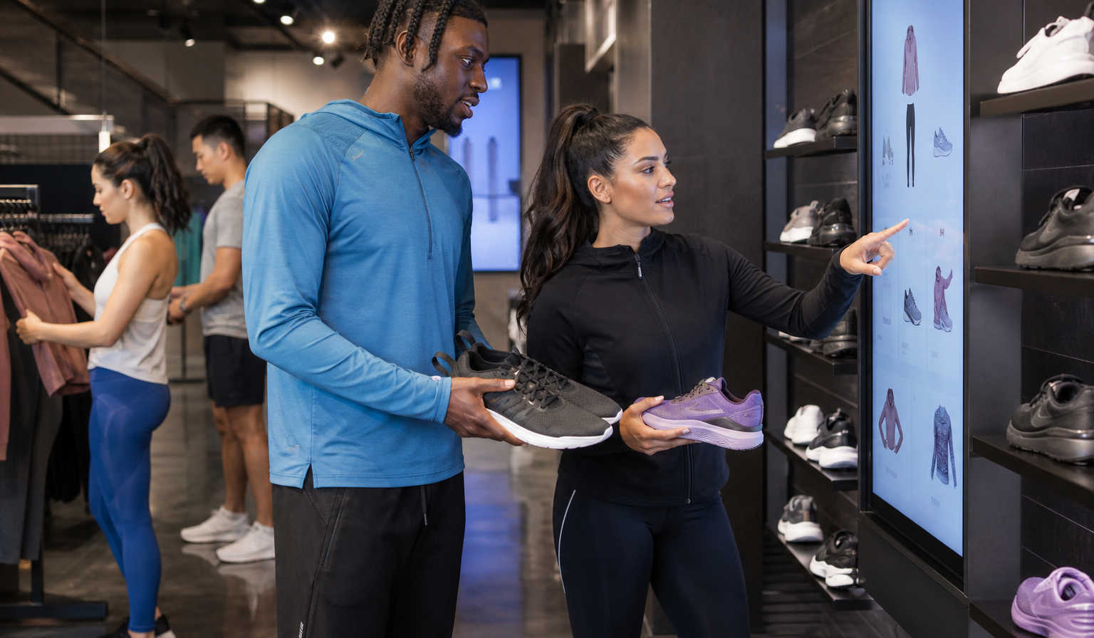Athletic and performance wear shoppers interacting with personalized AI recommendations on digital screens in a sleek sports retail environment, with running shoes, leggings and jackets displayed under dynamic lighting.